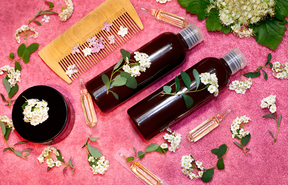 Vials with hair care product in fresh flowers on a pink towel.